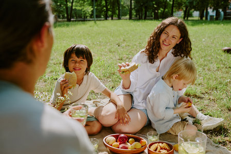 Caucasian woman sitting on grass with two children and young adult man having picnic, smiling and...