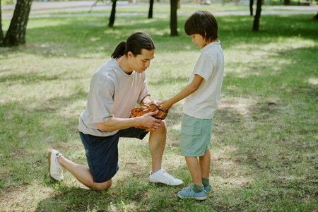 Young adult Asian man kneeling on grass putting baseball glove on biracial child boy standing in...