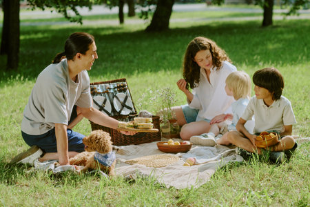 Middle aged woman sitting on picnic blanket with two children and her husband, sharing food outdoors...