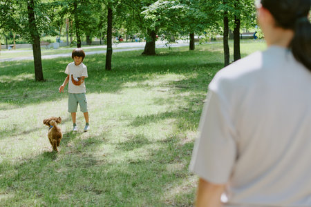 Biracial boy playing with small dog in green park while his parent with dark hair observing from foreground, sunlight filtering through trees, summer outdoor family activityの写真素材