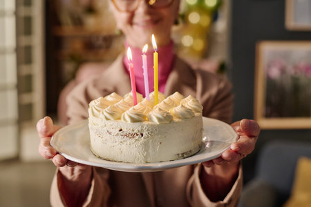 Senior woman holding birthday cake with three lit candles, smiling and presenting dessert toward...