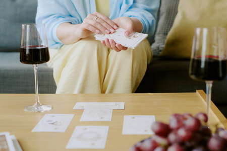 Woman shuffling tarot cards at table with wine glasses and grapes visible, arranging cards for...