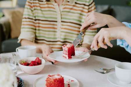Woman receiving slice of red cake from another guest, hands holding plate and utensils, sitting at table with strawberries and cupsの写真素材