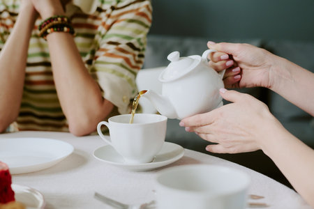 Woman pouring tea from teapot into cup while sitting at table with friends, both engaging in casual conversation during tea timeの写真素材