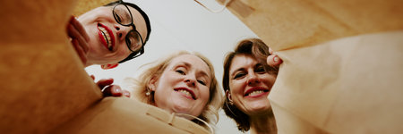 Three middle aged Caucasian women smiling and looking down into shopping bag, faces close together, showing joyful expressions, photographed from inside bag perspectiveの写真素材