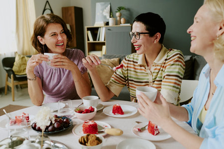 Three middle-aged Caucasian women sitting at table laughing and eating cake together, holding cups...