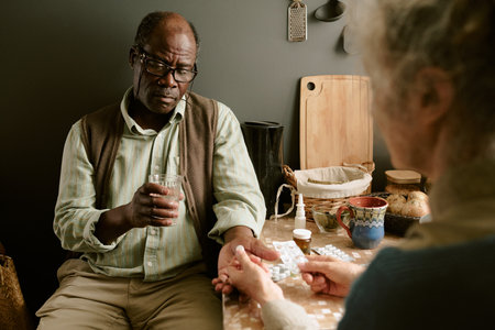Senior Black man sitting at table holding glass of water while senior Caucasian woman handing medication, both engaged in daily routine, kitchen background visibleの写真素材