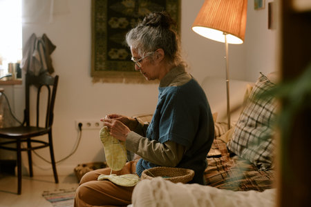 Senior Caucasian woman sitting on sofa knitting yellow garment, wearing glasses and focusing on...