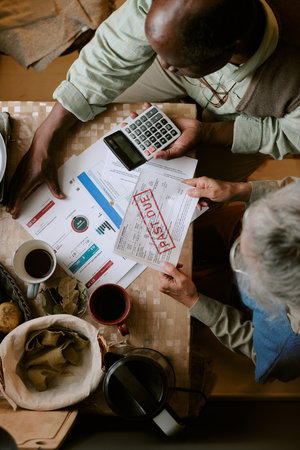 Senior Caucasian woman and senior Black man sitting at table reviewing overdue bills and financial...