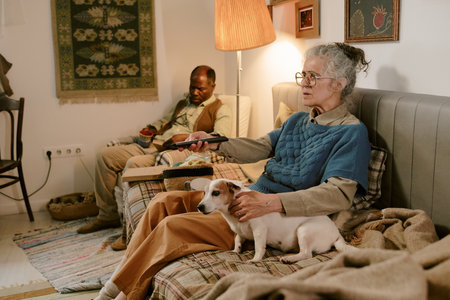 Senior woman sitting on sofa holding remote control with dog on lap, senior man sitting in...