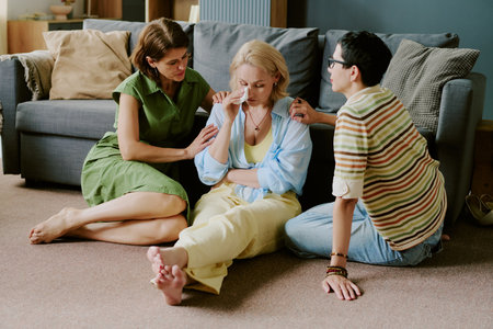 Middle aged Caucasian woman sitting on floor wiping tears while two women comforting her, all seated...