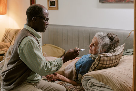 Senior Black man feeding senior Caucasian woman lying on couch, both showing signs of aging, man gently holding spoon near womans mouth, intimate caregiving moment in modest homeの写真素材