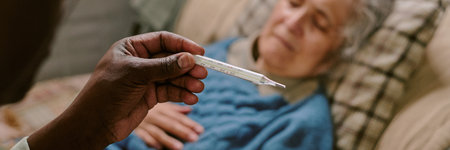 Senior Caucasian woman lying on couch with eyes closed, Black man holding thermometer in foreground checking temperature, illustrating elderly care and health monitoringの写真素材