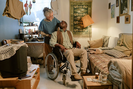 Senior Caucasian woman standing behind senior Black man sitting in wheelchair with disability, both...