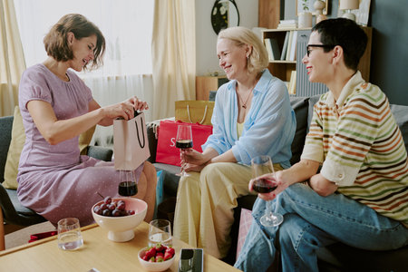 Three middle aged women sitting together on sofa smiling and talking while holding glasses of red...
