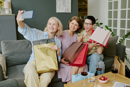 Three middle aged Caucasian women sitting on sofa smiling and holding shopping bags while taking...