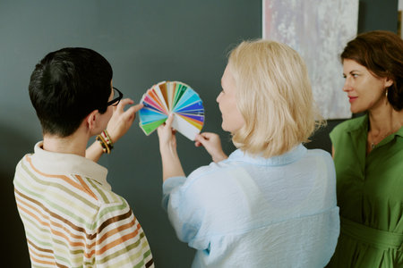 Three middle aged women standing together examining color swatch samples against dark wall,...
