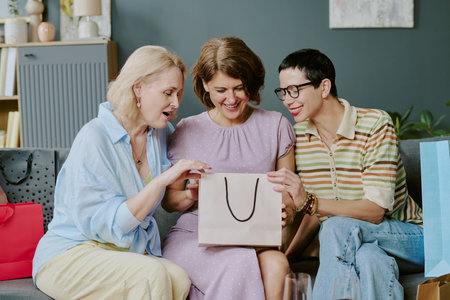 Three middle aged Caucasian women sitting together on sofa smiling and opening shopping bag, engaging in friendly conversation during leisure timeの写真素材