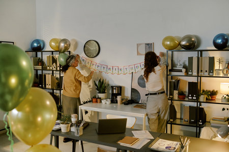 Two Caucasian women decorating office with balloons and hanging happy birthday banner on wall,...