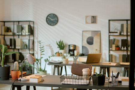 Modern office workspace featuring two desks with laptops, coffee cups, potted plants, stationery,...