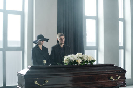 Caucasian young woman wearing sunglasses and hat standing beside Caucasian young man holding rose, both mourning near closed coffin with flowers in bright roomの写真素材