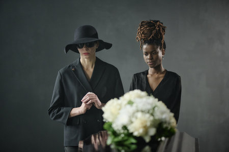 Caucasian middle aged woman wearing sunglasses and Black young adult woman standing beside coffin...