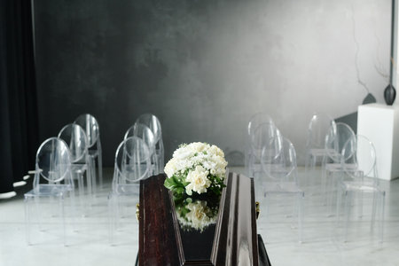 Closed wooden coffin with floral arrangement standing in front of empty transparent chairs in modern...