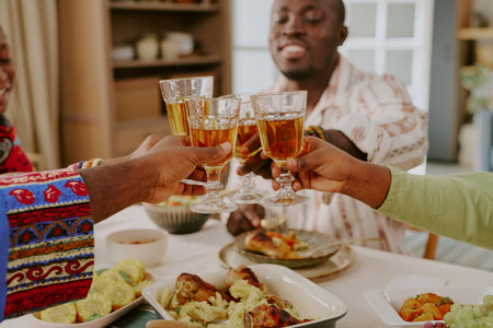 Group of Black adults clinking glasses while celebrating Kwanzaa at dining table, hands raised in toast, traditional food dishes visible in foreground, joyful expressions evidentの写真素材