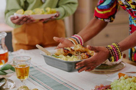 Black woman serving traditional food from casserole dish during Kwanzaa celebration, hands adorned with colorful beaded bracelets, standing beside another adult holding plate of vegetablesの写真素材
