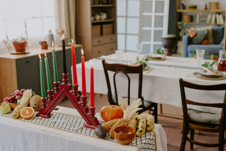 Kwanzaa kinara with seven lit candles standing on table surrounded by fruits, vegetables, corn and traditional decorations, dining table set for celebration visible in backgroundの写真素材
