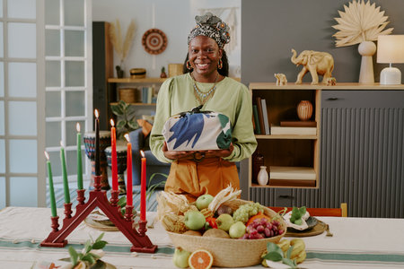 Portrait of young adult Black woman smiling while holding wrapped gift standing behind table with kinara, fruit basket, Kwanzaa decorations, celebrating traditional holiday indoorsの写真素材