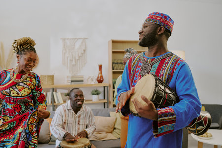 Black man wearing traditional clothing playing drum, while smiling Black woman and Black young adult...