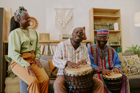 Three Black adults celebrating Kwanzaa, two men playing traditional drums while woman standing...