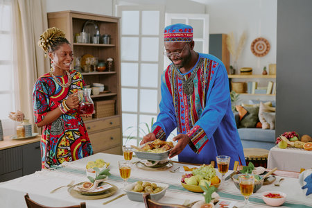 Middle aged Black man wearing traditional clothing serving food while smiling, standing beside young...
