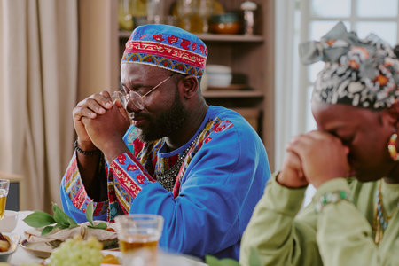 Black middle aged man and Black woman sitting at table clasping hands in prayer during Kwanzaa celebration, traditional attire and festive meal visible, eyes closed in reflectionの写真素材