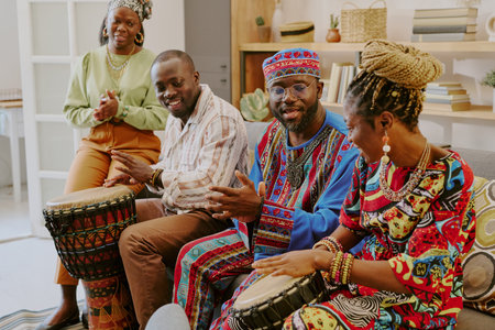 Group of Black adults celebrating Kwanzaa, sitting together on sofa, playing traditional drums, smiling and engaging in conversation, woman standing clapping hands nearbyの写真素材