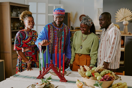 Group of Black adults standing around table celebrating Kwanzaa, man lighting kinara candles while others watching, traditional fruits and vegetables arranged in centerpieceの写真素材
