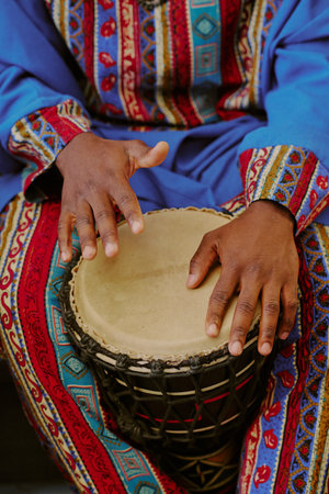 Black adult wearing traditional patterned clothing playing drum with hands, celebrating Kwanzaa,...
