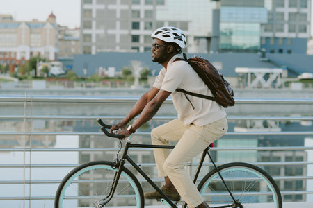 Young adult man riding bicycle along urban waterfront, wearing helmet and backpack, side view showing focused expression, modern city buildings in backgroundの写真素材