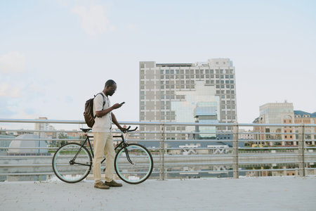 Young adult Black man standing outdoors next to bicycle using smartphone, wearing backpack, urban cityscape in background, appearing focused while interacting with deviceの写真素材