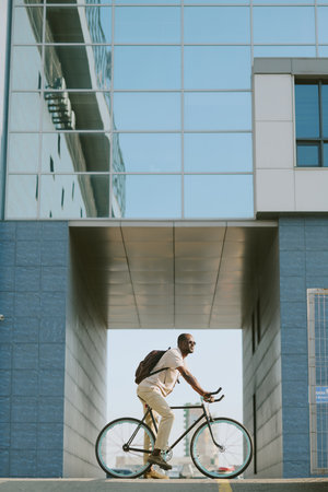 Young adult Black man riding bicycle outdoors in urban setting, wearing backpack and sunglasses, passing modern glass building, side profile visible, commuting or travelingの写真素材