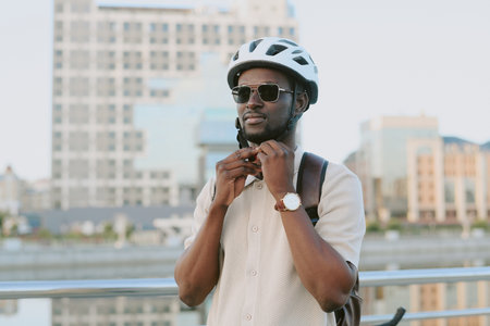 Young adult man standing outdoors adjusting bicycle helmet strap, wearing sunglasses and backpack, cityscape with modern buildings in background, looking away from cameraの写真素材