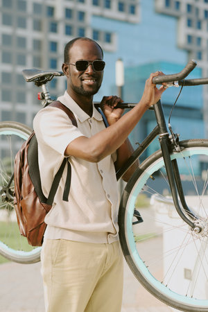 Portrait of young adult man wearing sunglasses smiling and carrying bicycle on shoulder outdoors in...