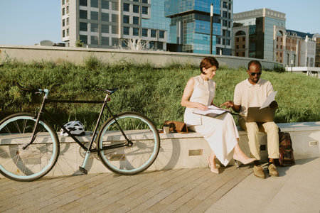Caucasian middle aged woman and Black middle aged man sitting outdoors on city bench collaborating with laptop and documents, bicycle parked nearby, both focused on workの写真素材