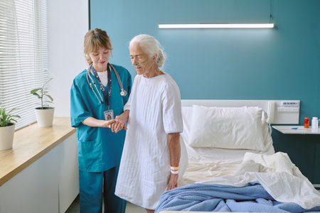 Caucasian female nurse assisting senior Caucasian woman with oxygen tube standing beside hospital bed in medical room, nurse holding patient hand and providing support during recoveryの写真素材