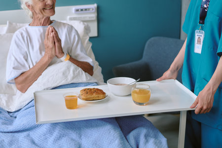Senior Caucasian woman sitting in hospital bed smiling with hands clasped while female nurse serving breakfast tray with juice, bread roll, and bowl of cereal in medical settingの写真素材