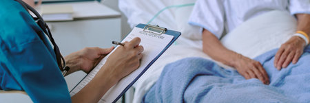 Woman wearing medical scrubs holding clipboard writing on medical history form while sitting beside senior patient, lying in hospital bed covered with blue blanketの写真素材