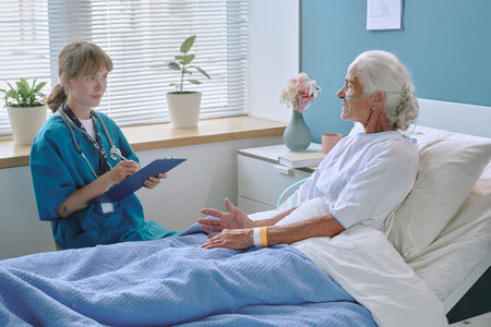 Senior Caucasian woman lying in hospital bed wearing oxygen tube, talking to young adult Caucasian...