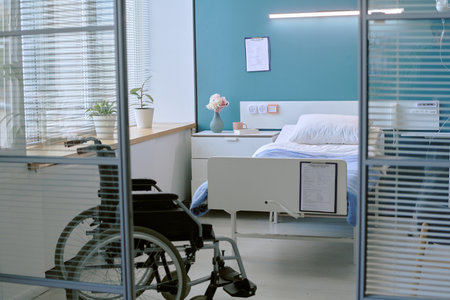 Empty hospital room featuring wheelchair with disability positioned near bed, medical equipment mounted on wall, potted plants on windowsill, clipboard attached to bed frameの写真素材