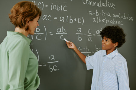 Black teenage boy explaining math equation on chalkboard while middle aged Caucasian woman listening attentively in classroom setting, both standing and interacting during lessonの写真素材